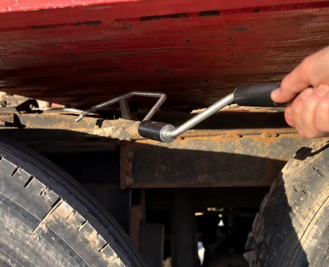 A driver reaches under a trailer between the tires with the metal tool to pull the fifth wheel king pin release